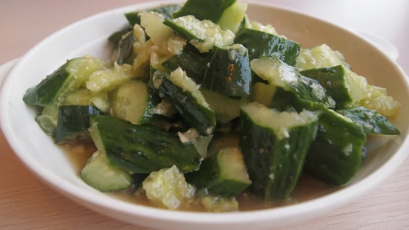 Sichuan Smashed Cucumber Salad Recipe: Texture, Dressing, and Serving Steps 3 Irregular smashed cucumber pieces with chopped garlic and a light dressing on a white plate