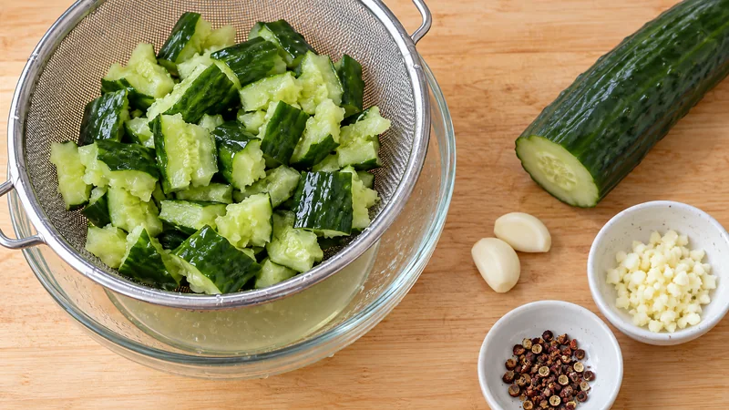 Cold Mixed Cucumber: Sichuan Recipe and Home Cooking Tips 2 Smashed raw cucumber draining in a colander over a glass bowl